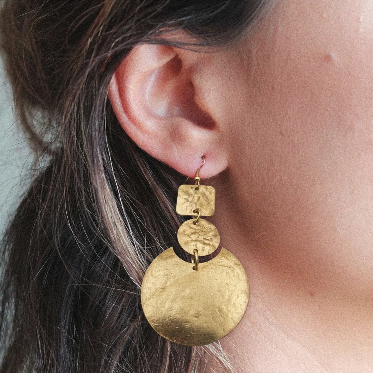 Woman wearing the Celestial Harmony Statement Earrings, handcrafted in brass with layered geometric discs – square, round, and crescent shapes – styled in a modern boho lifestyle setting.