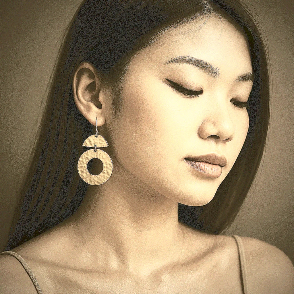 Model wearing Geo Sunburst Brass Earrings – handmade geometric earrings with bold hammered brass texture, shown in a soft Mediterranean-Balinese lifestyle setting.