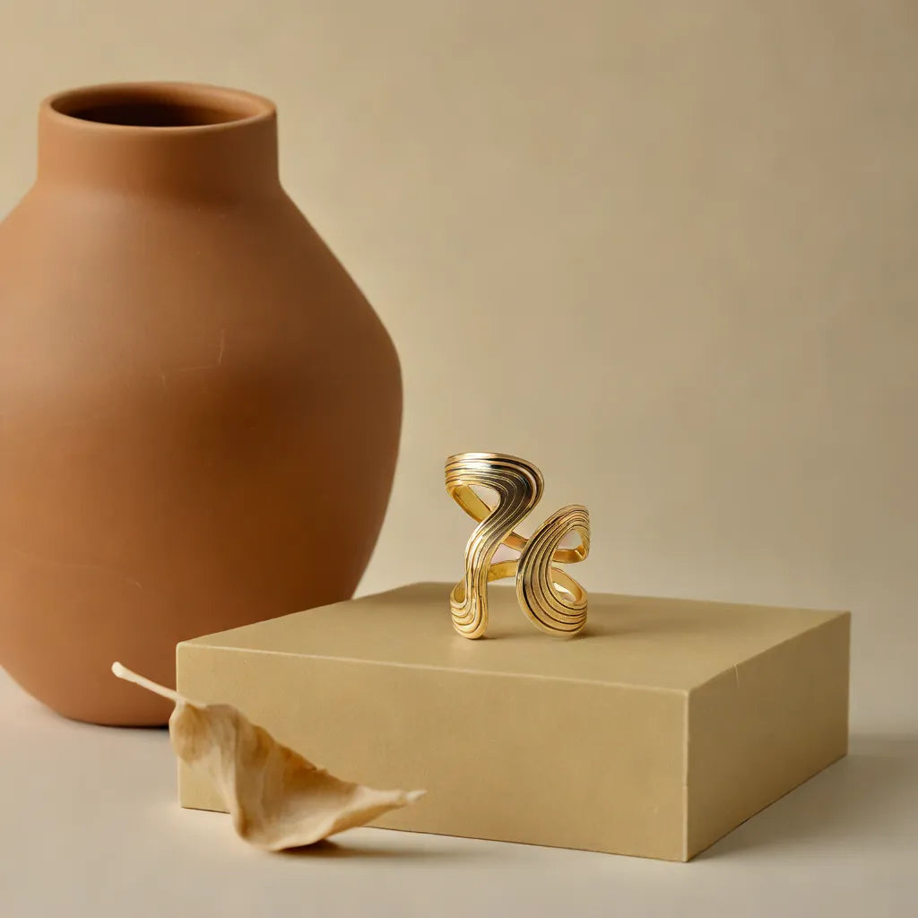 Handcrafted gold statement ring with flowing sculptural lines, placed on a neutral pedestal beside a terracotta vase and dried leaf — captured in soft Mediterranean light for Camins Fashion's artisanal boho collection.