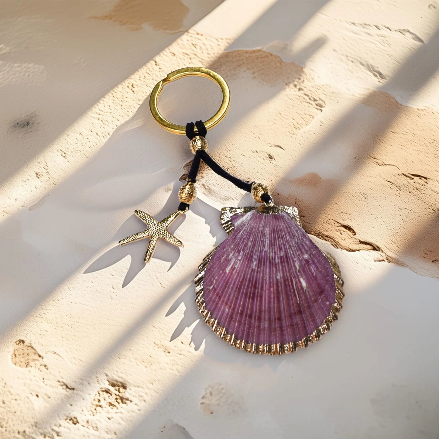Purple scallop shell keychain with 18K gold-plated trim and starfish charm, styled on a sunlit Mediterranean-textured surface — boho coastal accessory from Camins Fashion.