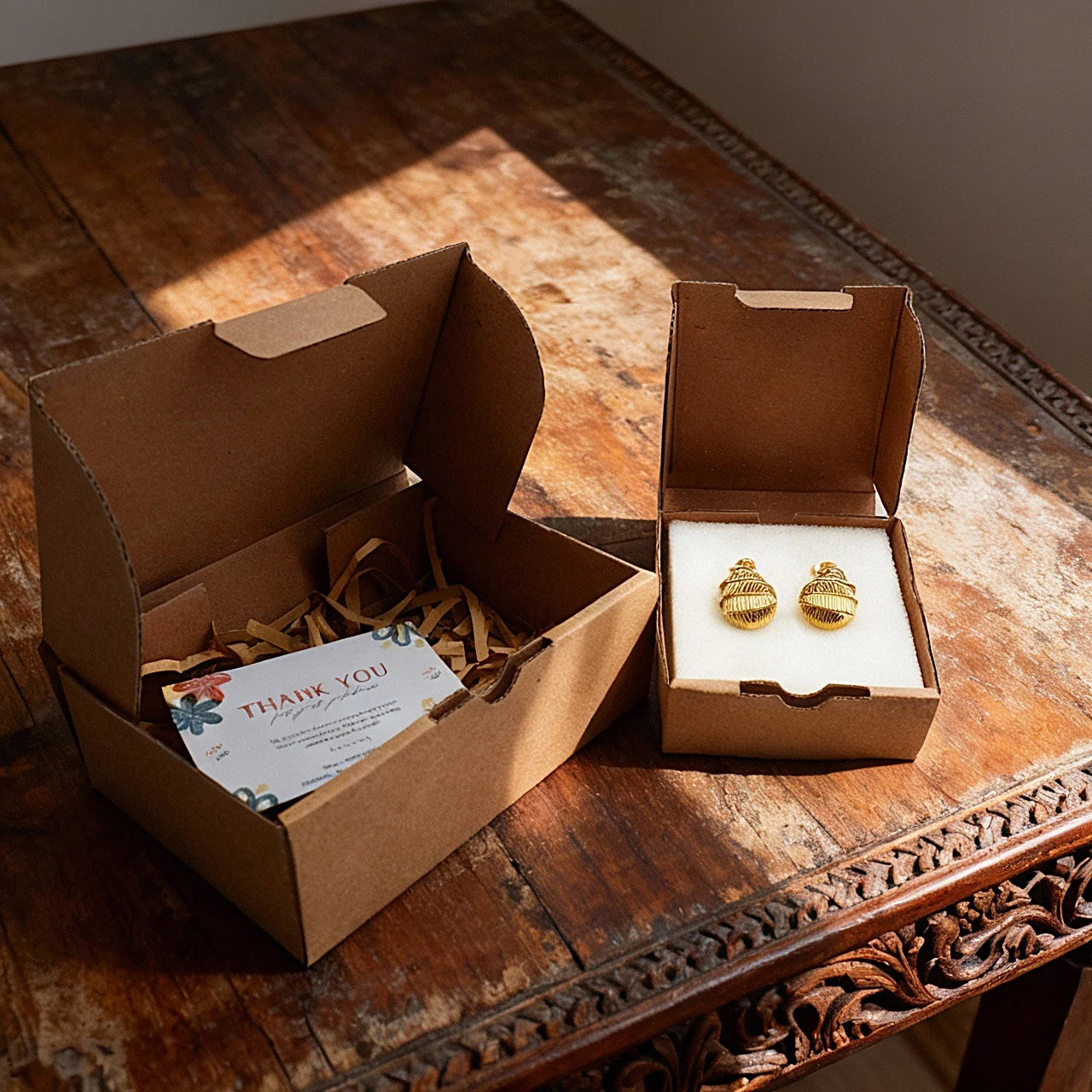 Handmade Camins jewelry gift packaging with gold-plated earrings on a carved Balinese artisan table in natural sunlight.