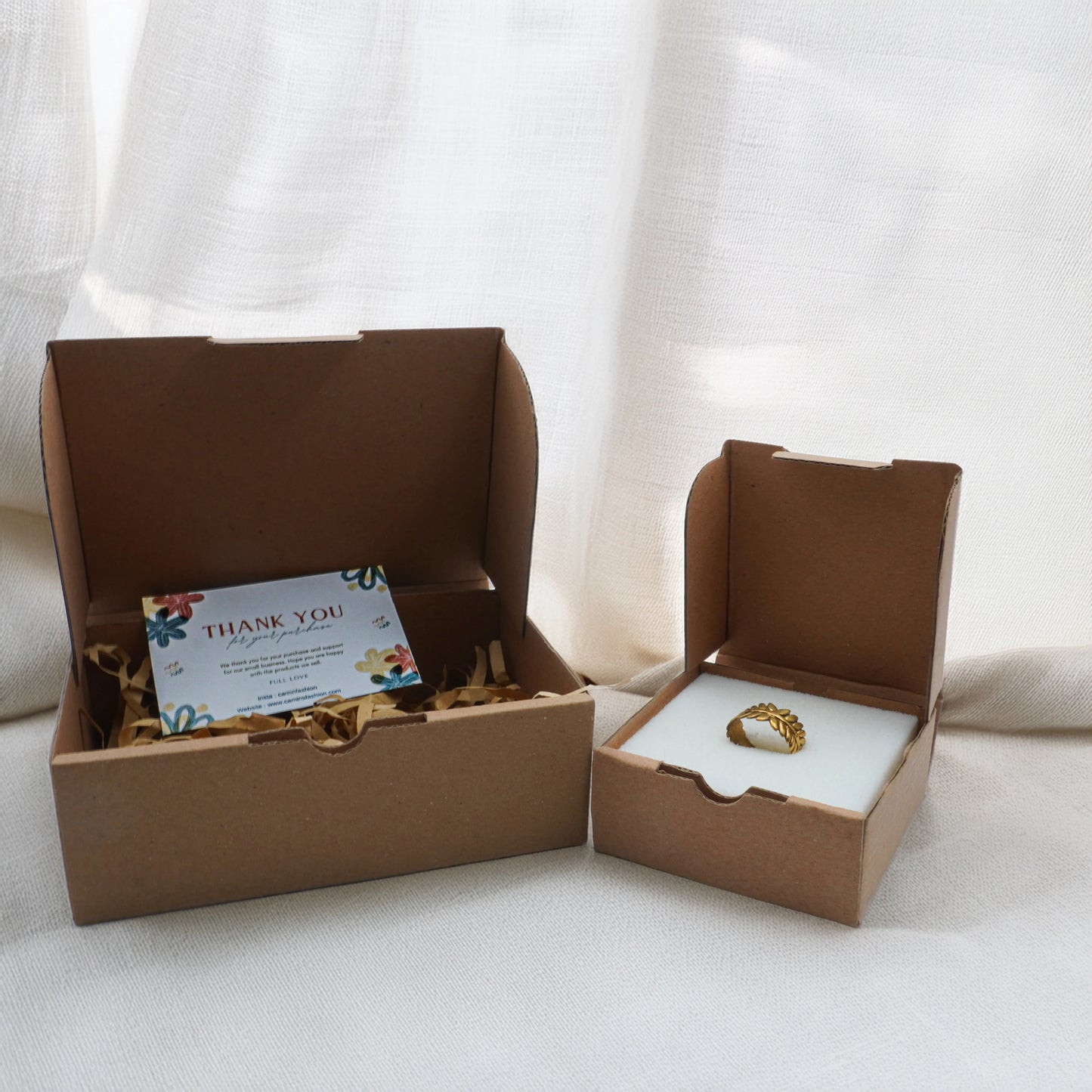 Camins Fashion eco-friendly jewelry packaging with thank-you card and gold-tone ring presented in sustainable kraft boxes on a soft cream fabric backdrop.
