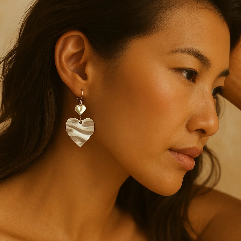 Elegant woman wearing silver heart-shaped earrings with a pearl accent, styled in a modern romantic look. Close-up profile shot with soft lighting, highlighting the sculptural design and minimalist Mediterranean aesthetic.