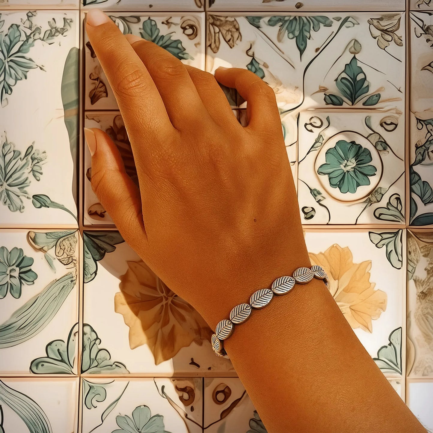 Close-up of a woman’s hand wearing the Serene Leaf Bracelet, a minimalist silver-toned bracelet with leaf-shaped beads, gently lit by natural sunlight. The hand rests on Mediterranean-style floral ceramic tiles, evoking calm, elegance, and nature-inspired design.