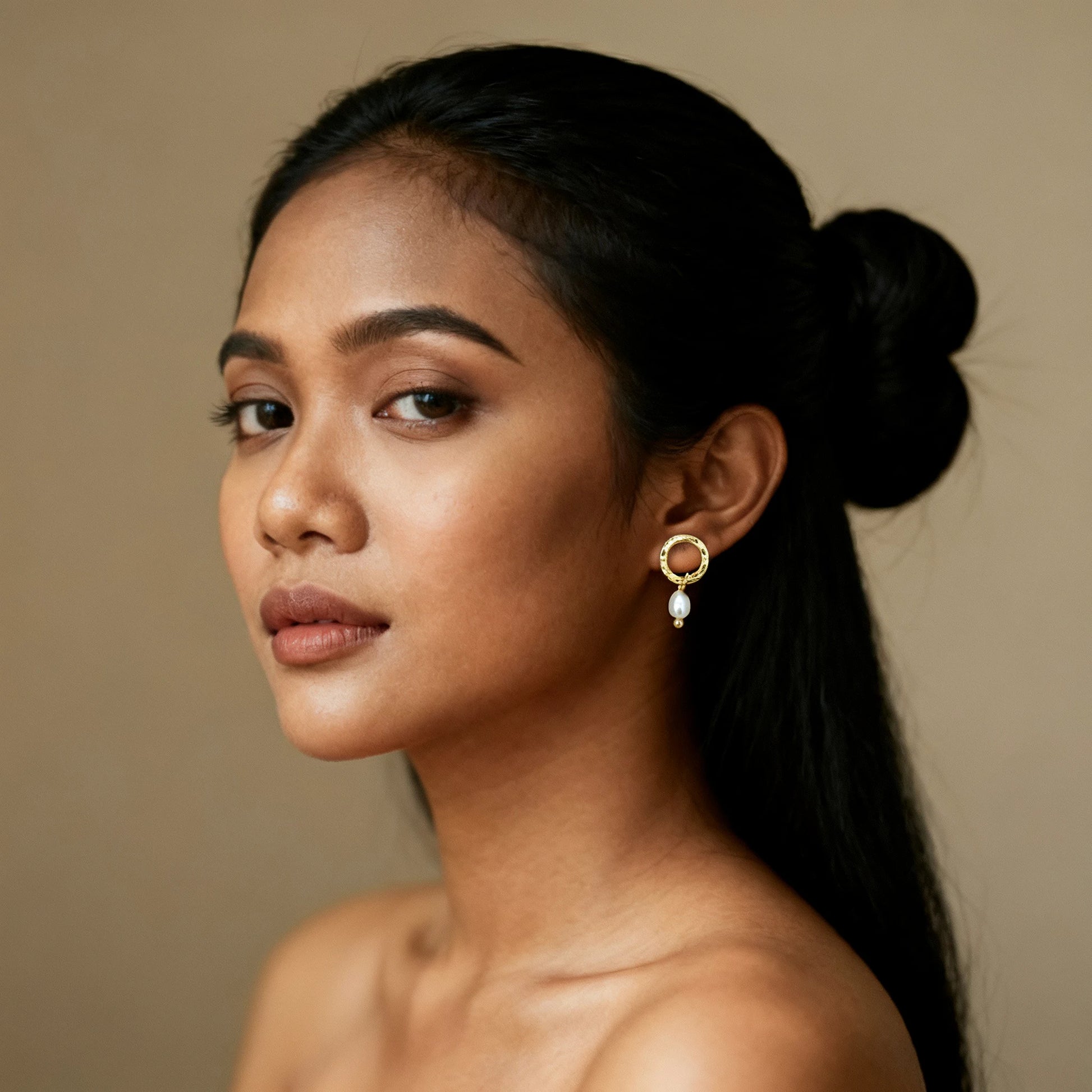 Portrait of a woman wearing Camins Golden Pearl Earrings, featuring a circular 18K gold-plated hoop with a single pearl drop, photographed against a warm neutral background.