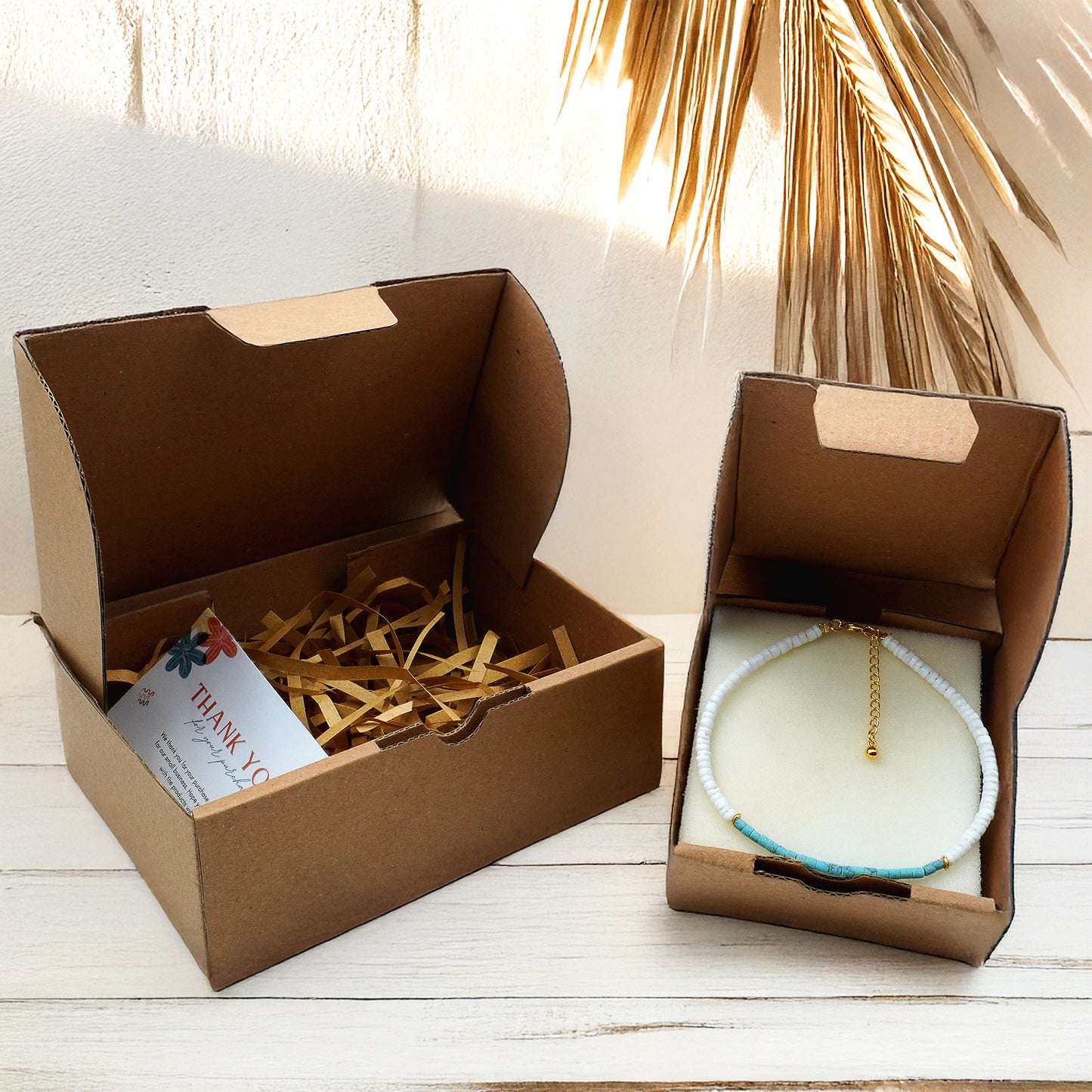 A natural brown kraft box packaging for handmade anklets, featuring eco-friendly shredded filler and a thank-you card on the left. On the right, a white and turquoise bead anklet with gold clasp is neatly displayed inside a box, styled with soft natural light and dried palm leaves in the background.