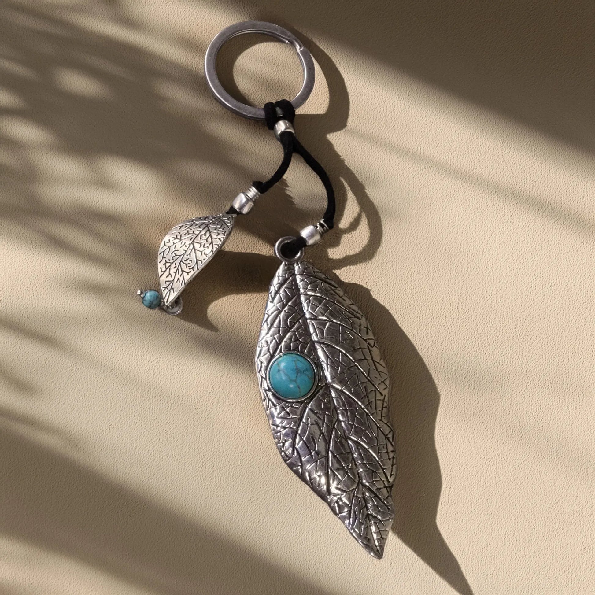 Artisan silver leaf keychain with etched botanical texture and turquoise stone center, hanging on a black cord under dappled sunlight — a handmade boho-chic accessory from Camins Fashion.