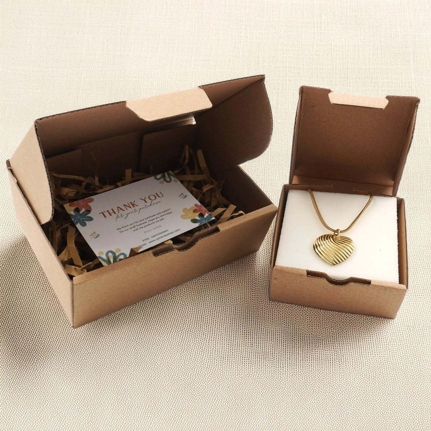 Eco-friendly jewelry packaging from Camins Fashion featuring a gold heart pendant necklace in a kraft gift box with thank you card and shredded paper, styled on a linen-textured background.