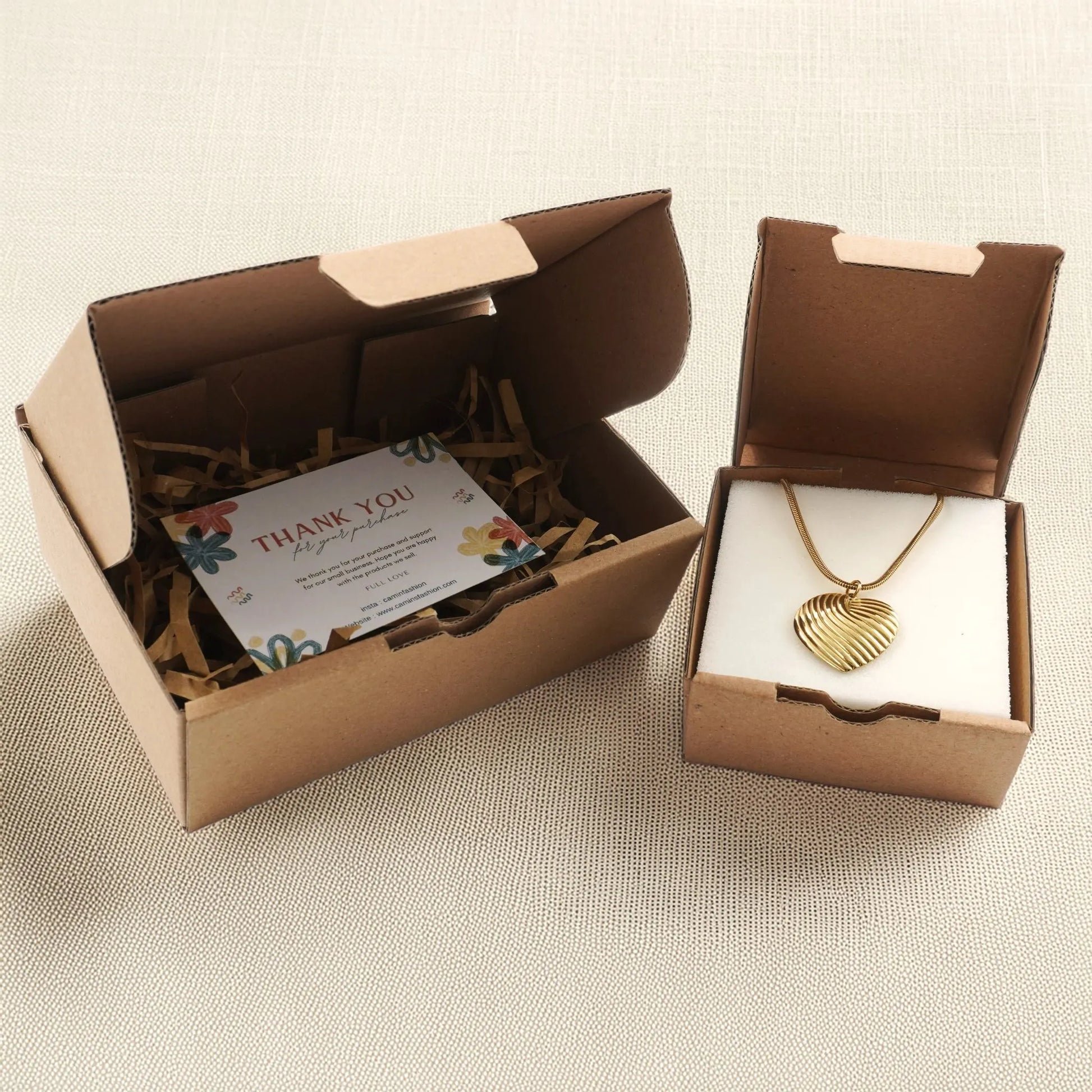 Eco-friendly jewelry packaging from Camins Fashion featuring a gold heart pendant necklace in a kraft gift box with thank you card and shredded paper, styled on a linen-textured background.