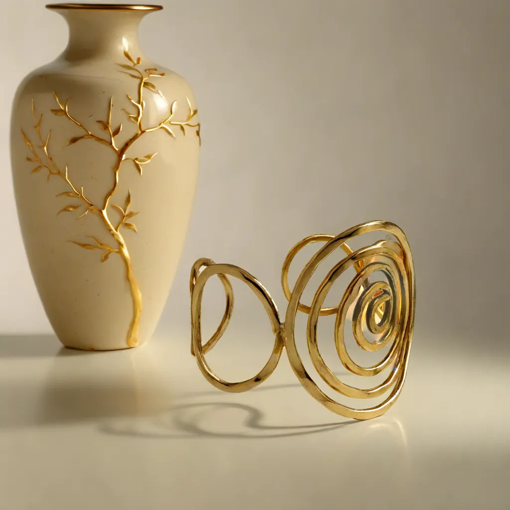 Sculptural gold spiral cuff bracelet placed beside a cream ceramic vase with golden botanical detailing, captured in warm Mediterranean light — a boho-chic statement piece reflecting Camins Fashion’s artisanal and minimalist elegance